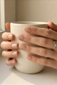 Simple, elegant nude-colored nails, hands holding a relaxing coffee cup in the gentle morning sunlight.