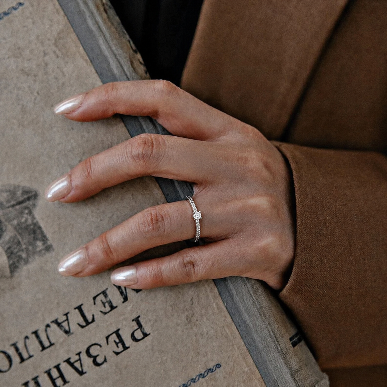 A hand with polished, almond-shaped nails holds a book, wearing a brown wool coat and a delicate ring.