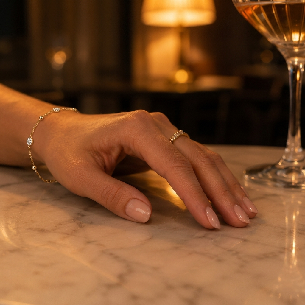 A close-up of a well-manicured hand with a high-shine top coat on nude nails, reflecting warm evening ambient light on a marble surface.