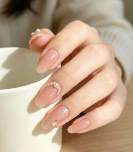 Glossy nude pink almond-shaped nails with delicate pearl embellishments under soft light.