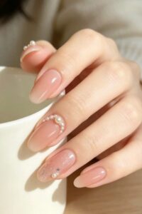 Glossy nude pink almond-shaped nails with delicate pearl embellishments under soft light.