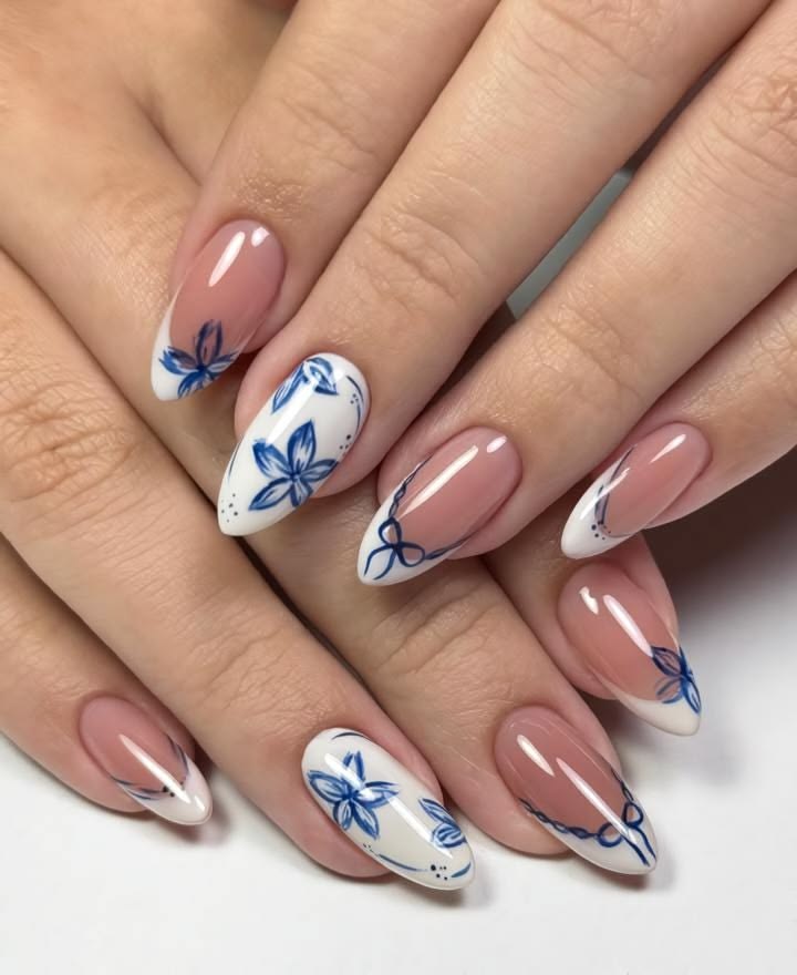 Elegant almond nails featuring delicate blue porcelain-inspired floral art on a glossy nude base for a timeless feminine look