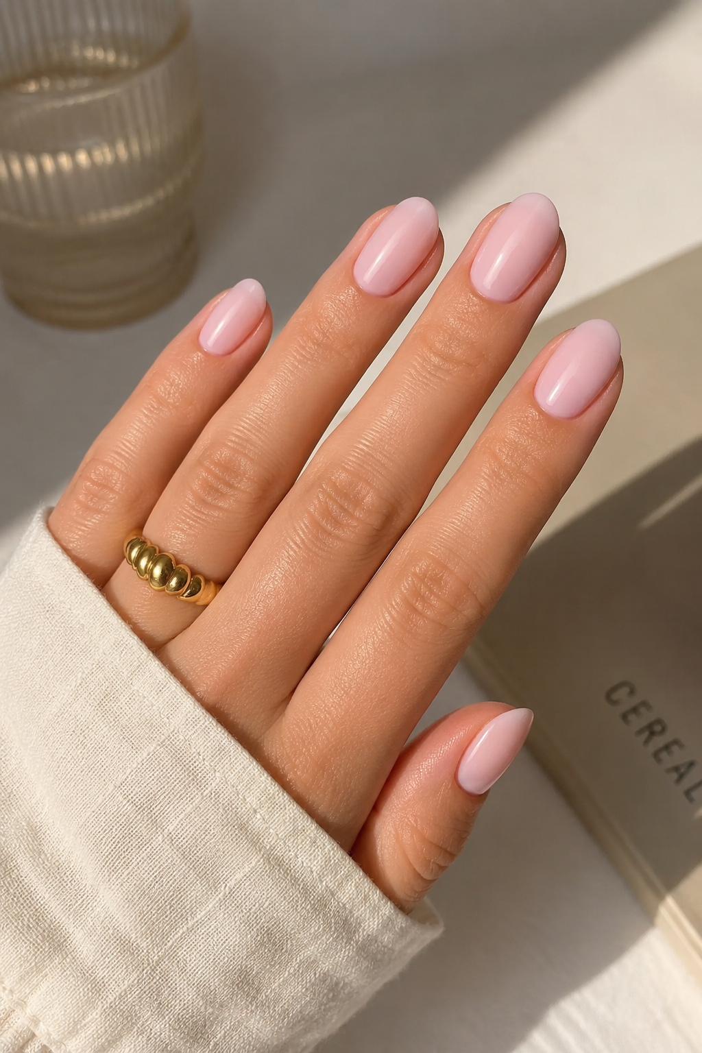 Milky pink summer nails on oval-shaped hands with a glossy finish, styled with a gold ring.