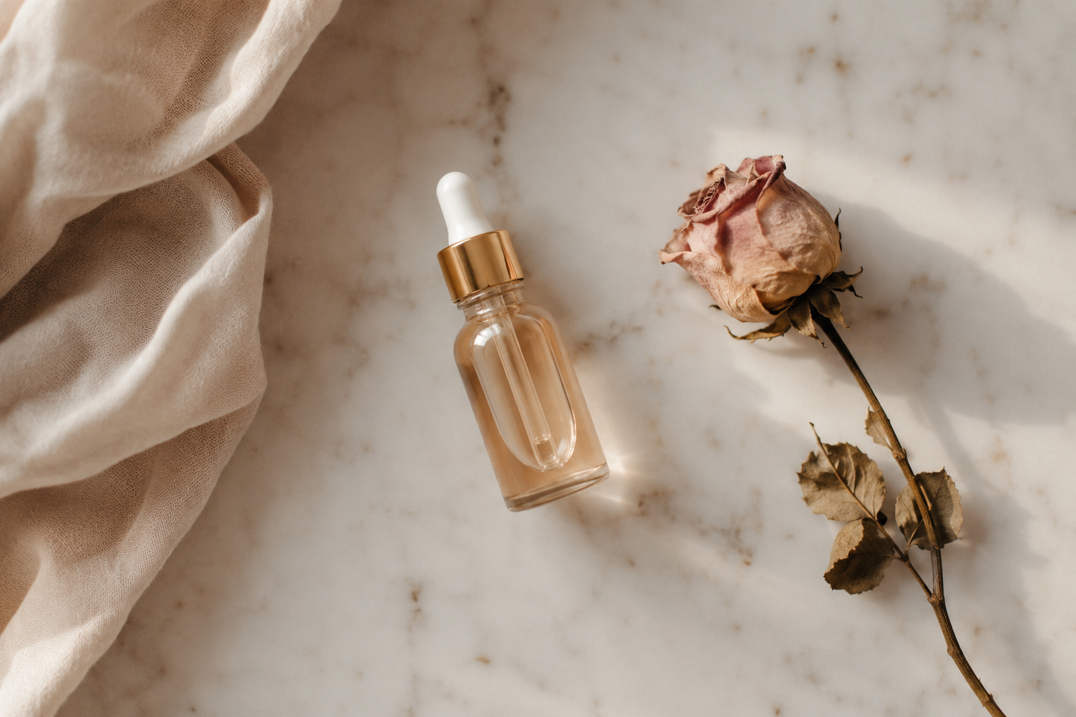 Minimalist flatlay of a glass cuticle oil dropper bottle on a marble surface with softly blurred hands in the background 